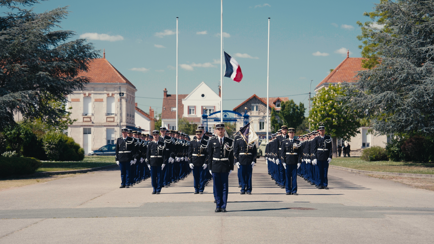 French military parade with uniformed officers marching in formation under a French flag at half-mast on a sunny day.