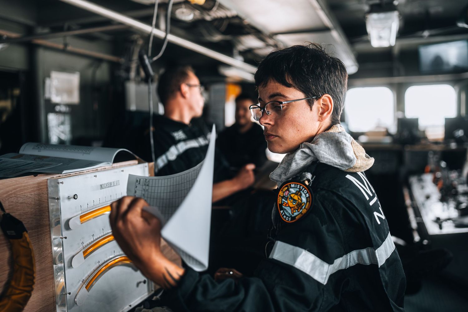 Young naval officer in uniform reviewing navigation charts on ship bridge.