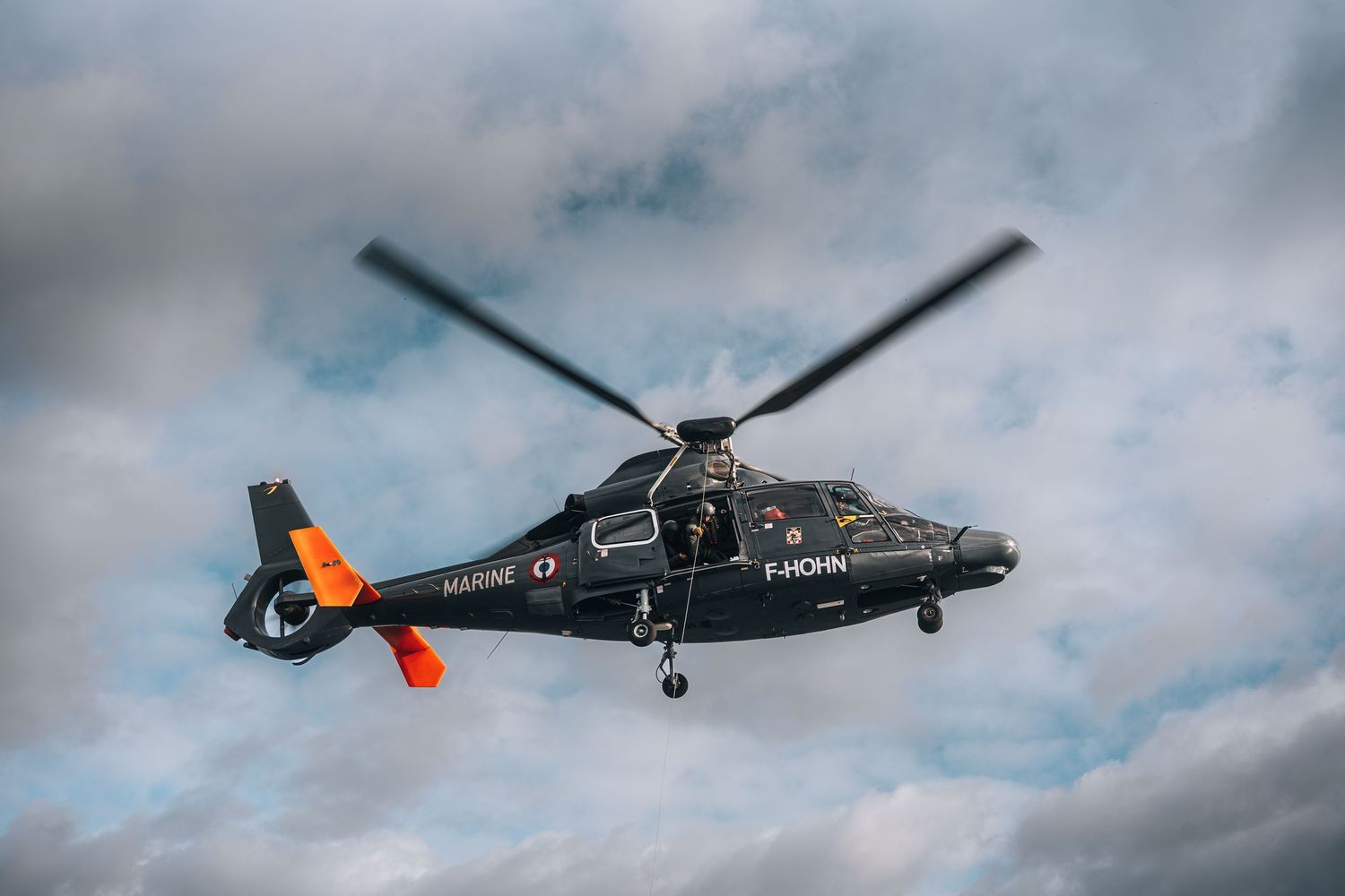 French Marine Nationale black military helicopter F-HOHN flies against cloudy sky.