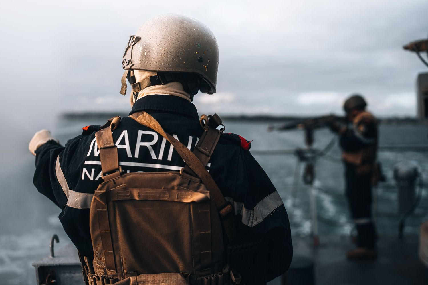 French Navy Marine standing on warship deck in tactical gear overlooking misty ocean waters.