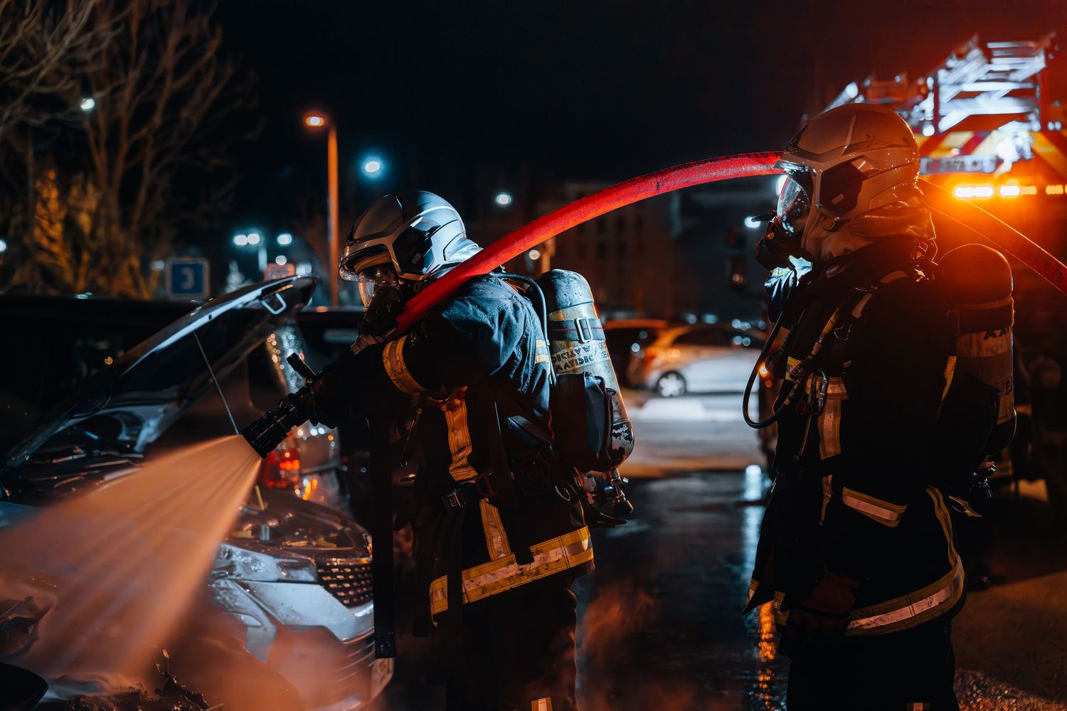 Firefighters in full gear use a red hose to extinguish a vehicle fire in a dark parking lot at night.