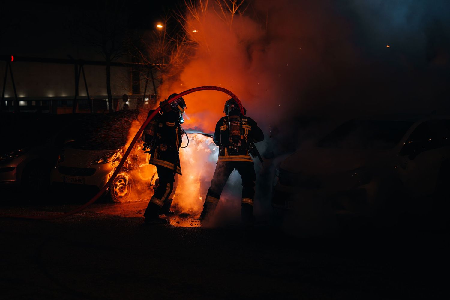 Two firefighters battle a blazing car fire at night, surrounded by bright orange flames and smoke.