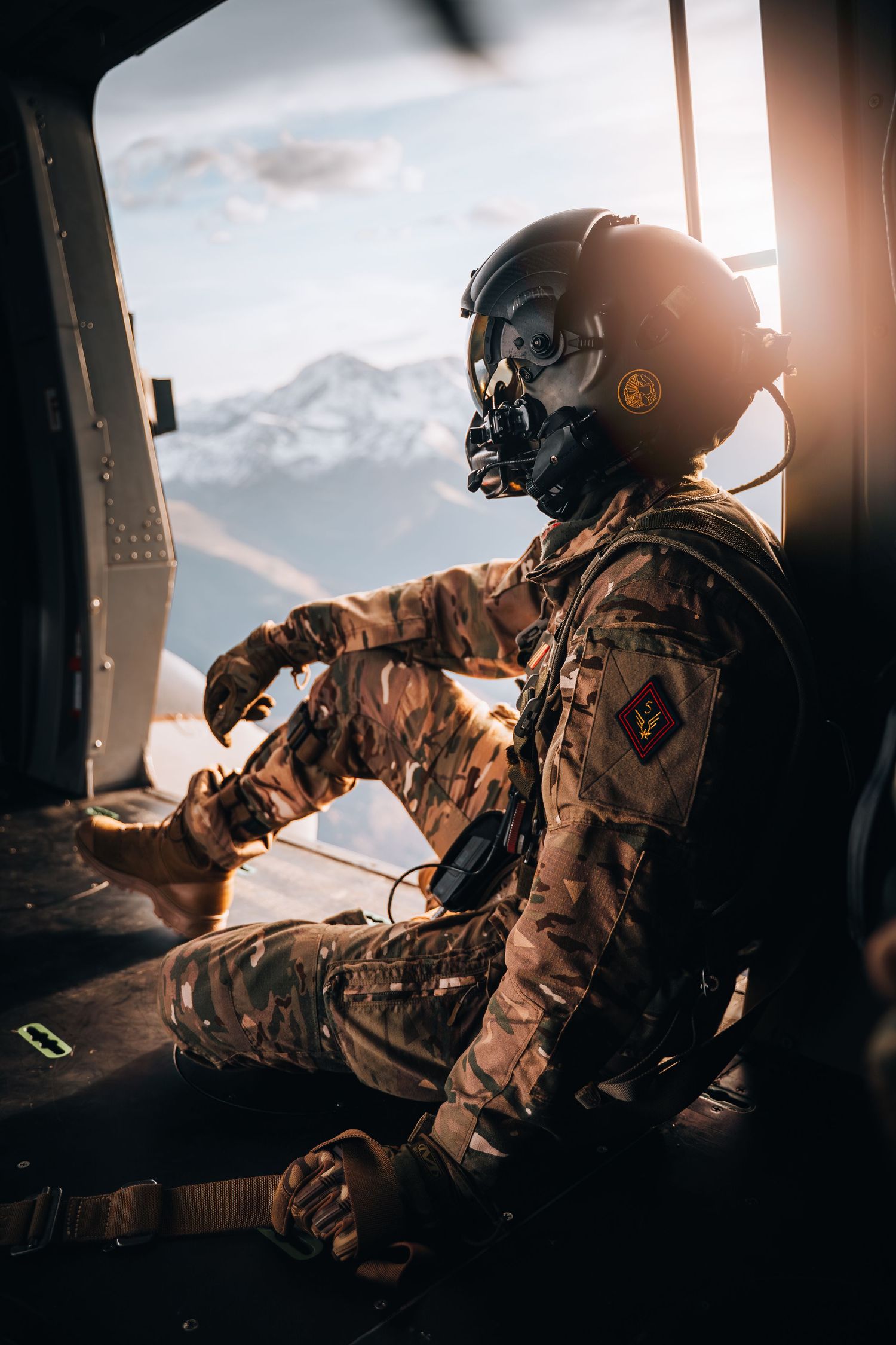 Armed soldier in full tactical gear sits in open helicopter door with mountain views behind.