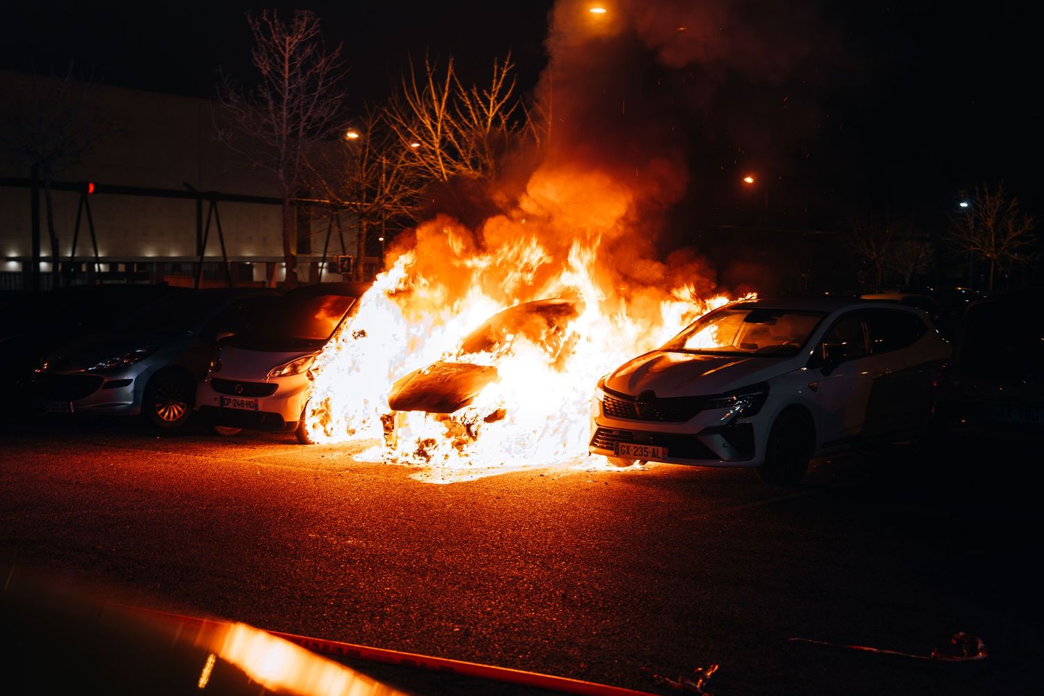 Multiple cars engulfed in intense orange flames burn in a parking lot during the night.