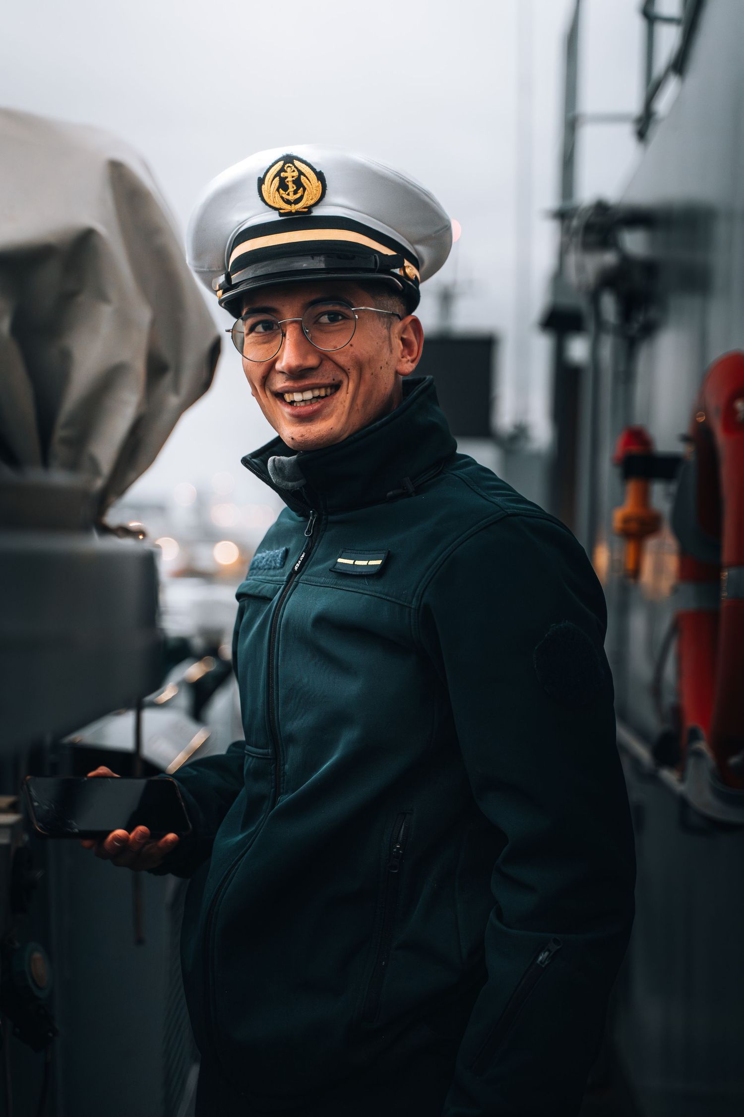 Smiling naval officer in white captain hat and dark uniform standing on ship deck in overcast weather.