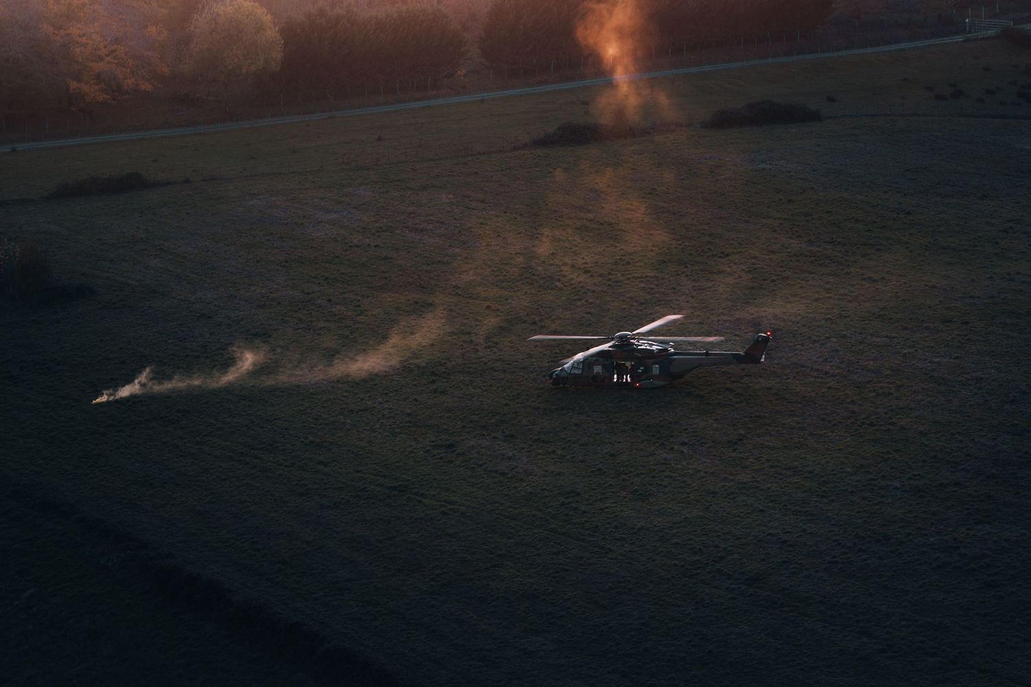 Military helicopter on a field at dusk with smoke rising from nearby ground fires.