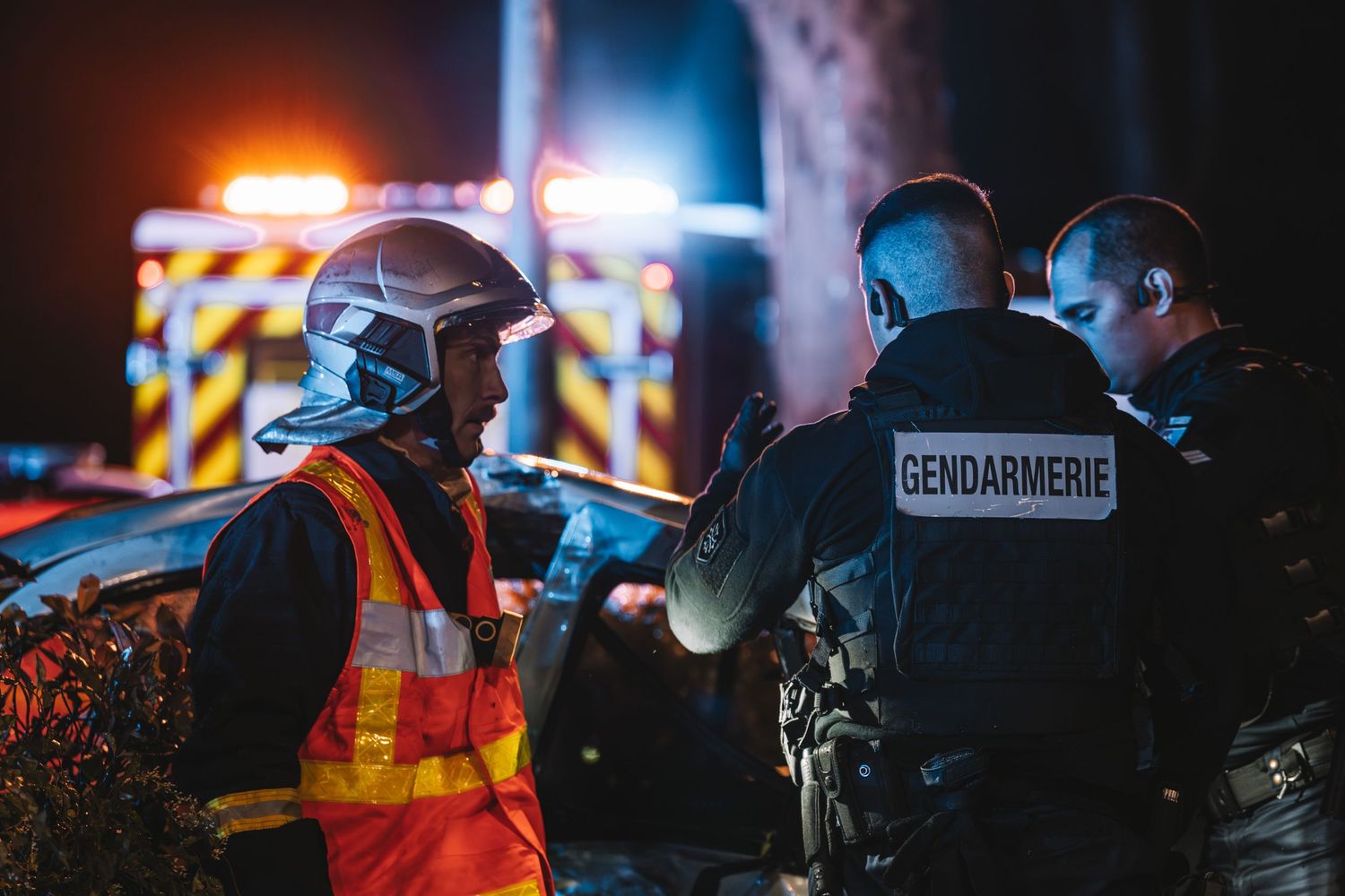A firefighter and two gendarmes coordinate at a nighttime emergency scene with fire trucks in the background.