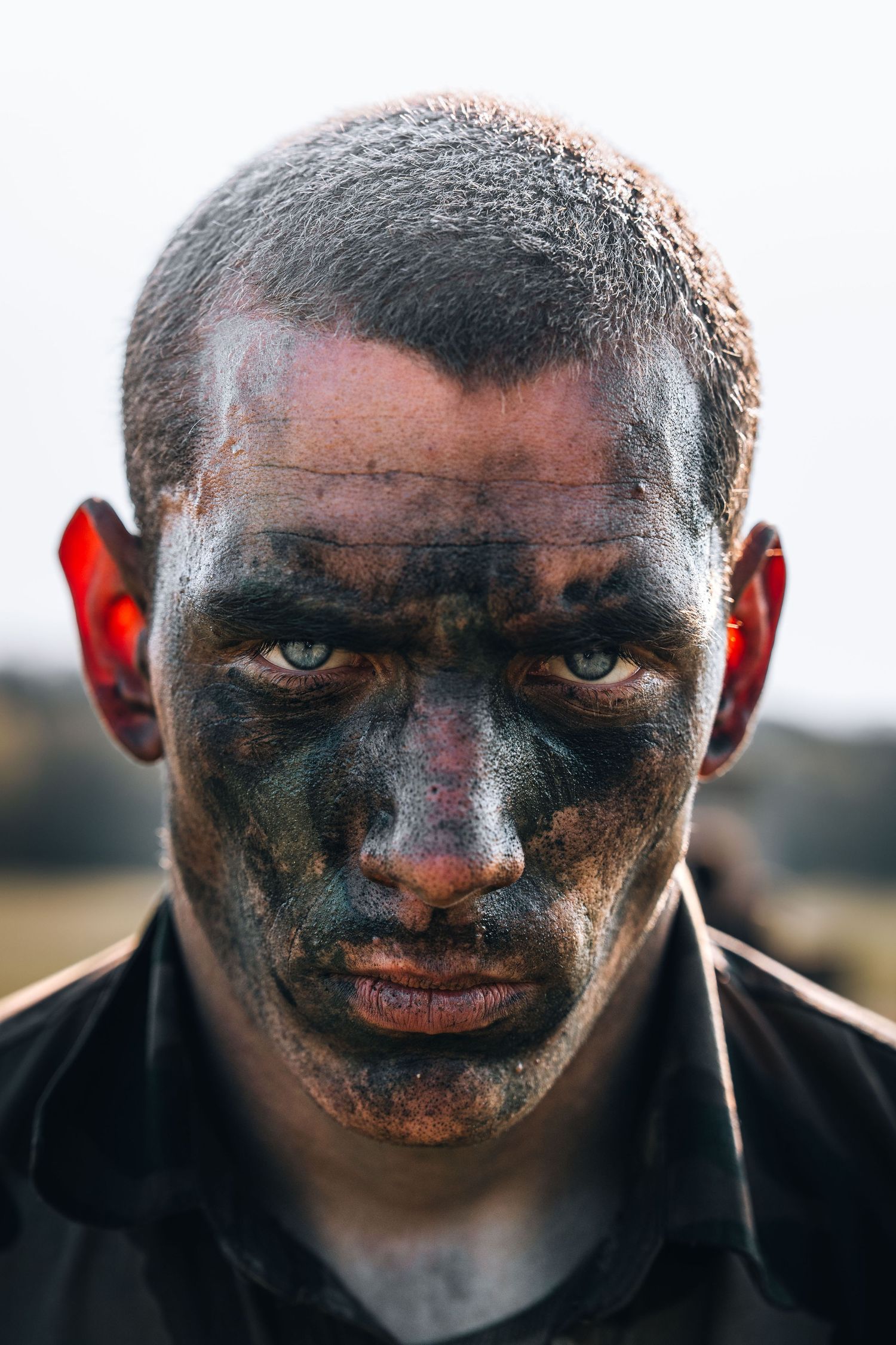 Close-up portrait of a man with camouflage face paint and intense blue eyes, wearing red ear defenders outdoors.