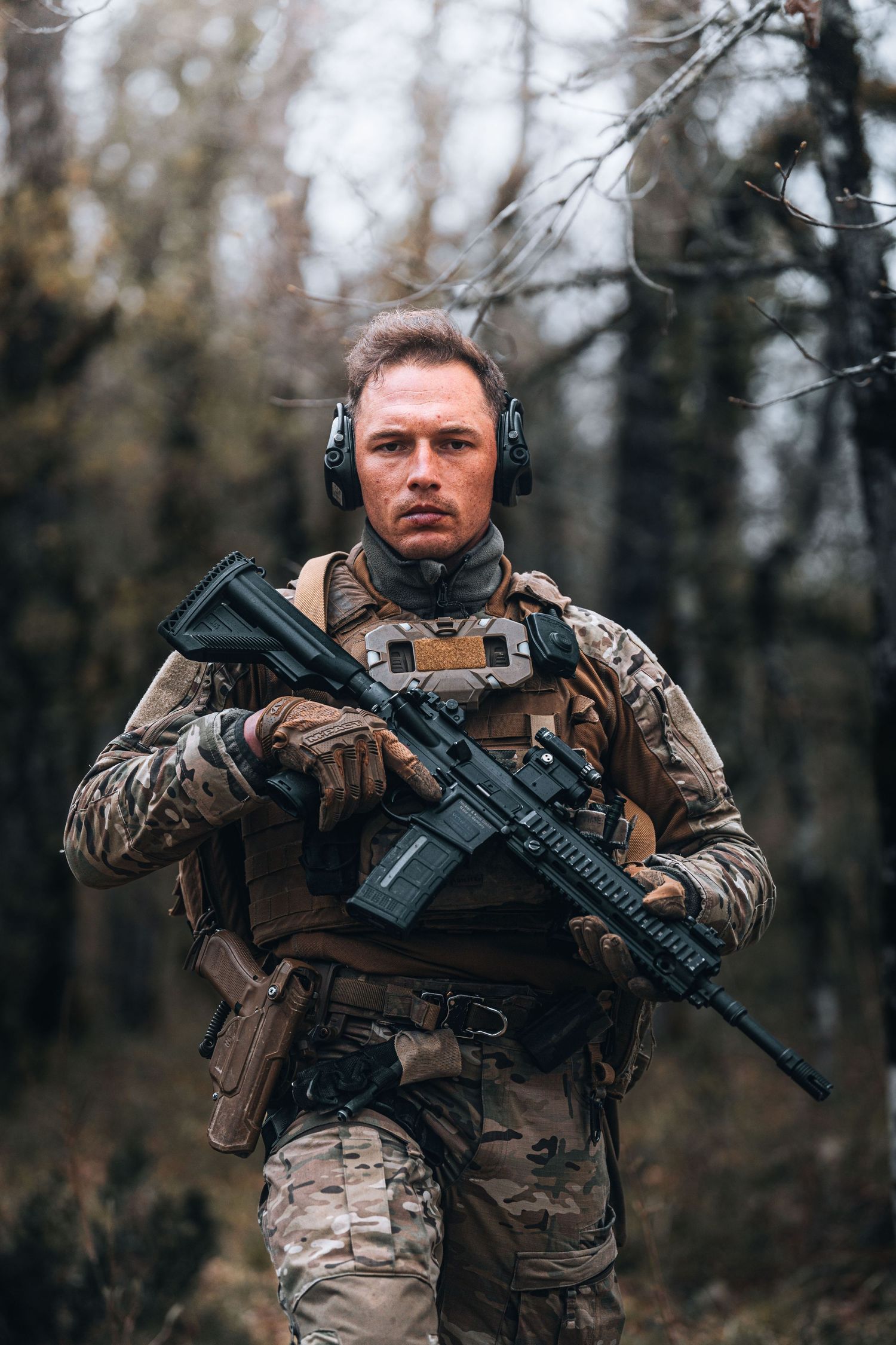 Armed soldier in camouflage gear and tactical equipment standing in a forest holding a rifle.