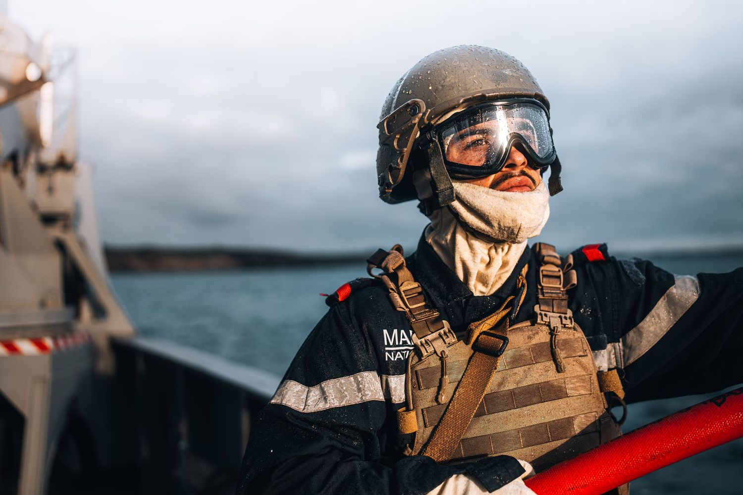 French Marine soldier in helmet and tactical vest stands on naval vessel with overcast sky.