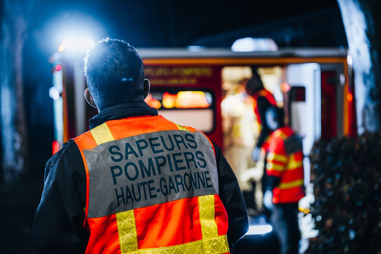A Sapeurs Pompiers Haute-Garonne firefighter in an orange safety vest stands near a fire truck at night.