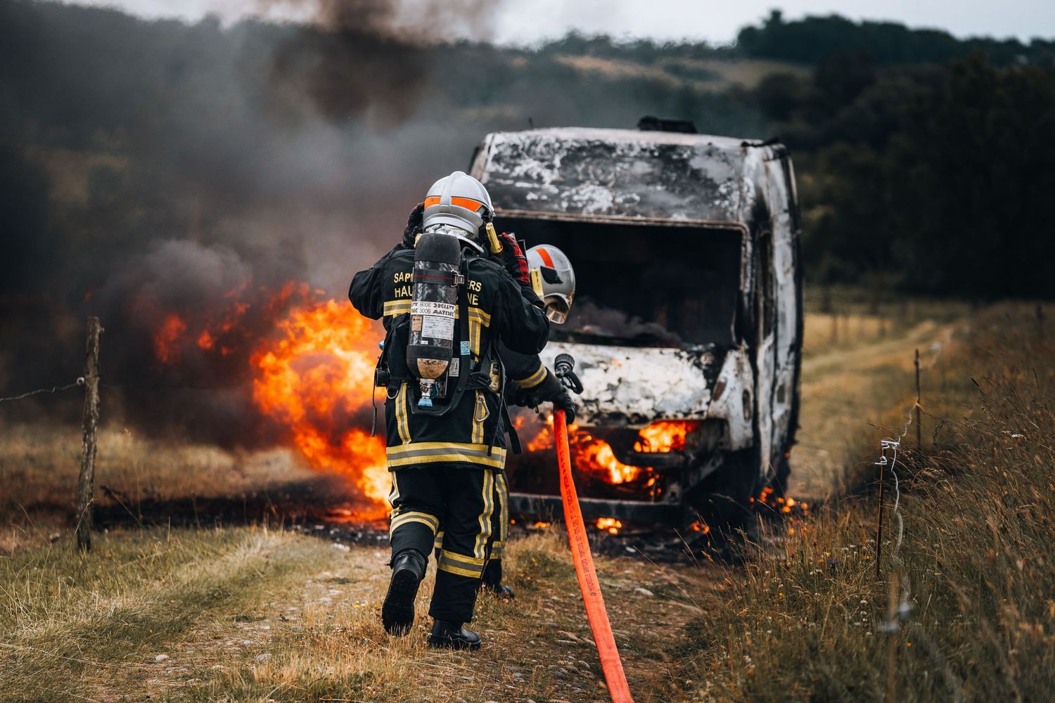 Firefighter in protective gear holding hose battling burning vehicle in rural field.