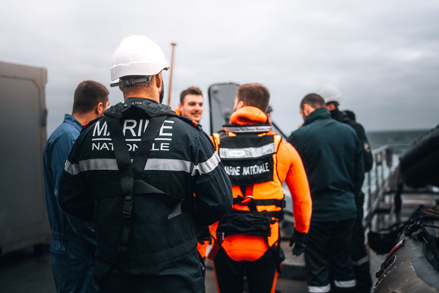 Marine Nationale sailors in black uniforms assisting person in orange life vest on ship deck.