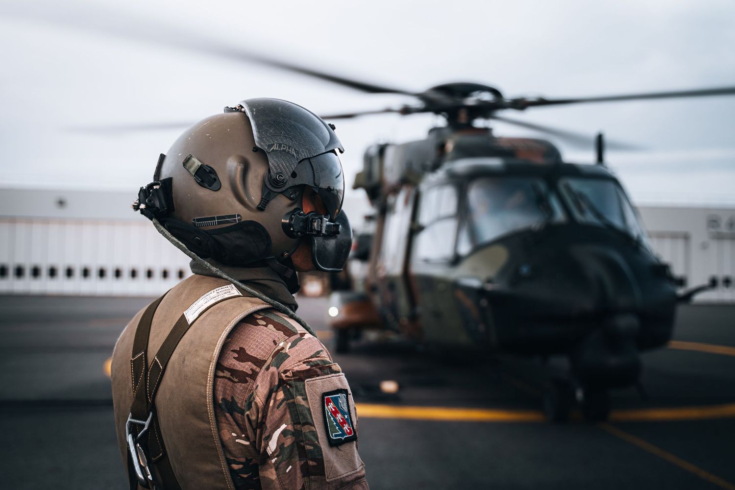 Military soldier in combat gear stands before a helicopter on a tarmac, wearing a tactical helmet and camouflage uniform.
