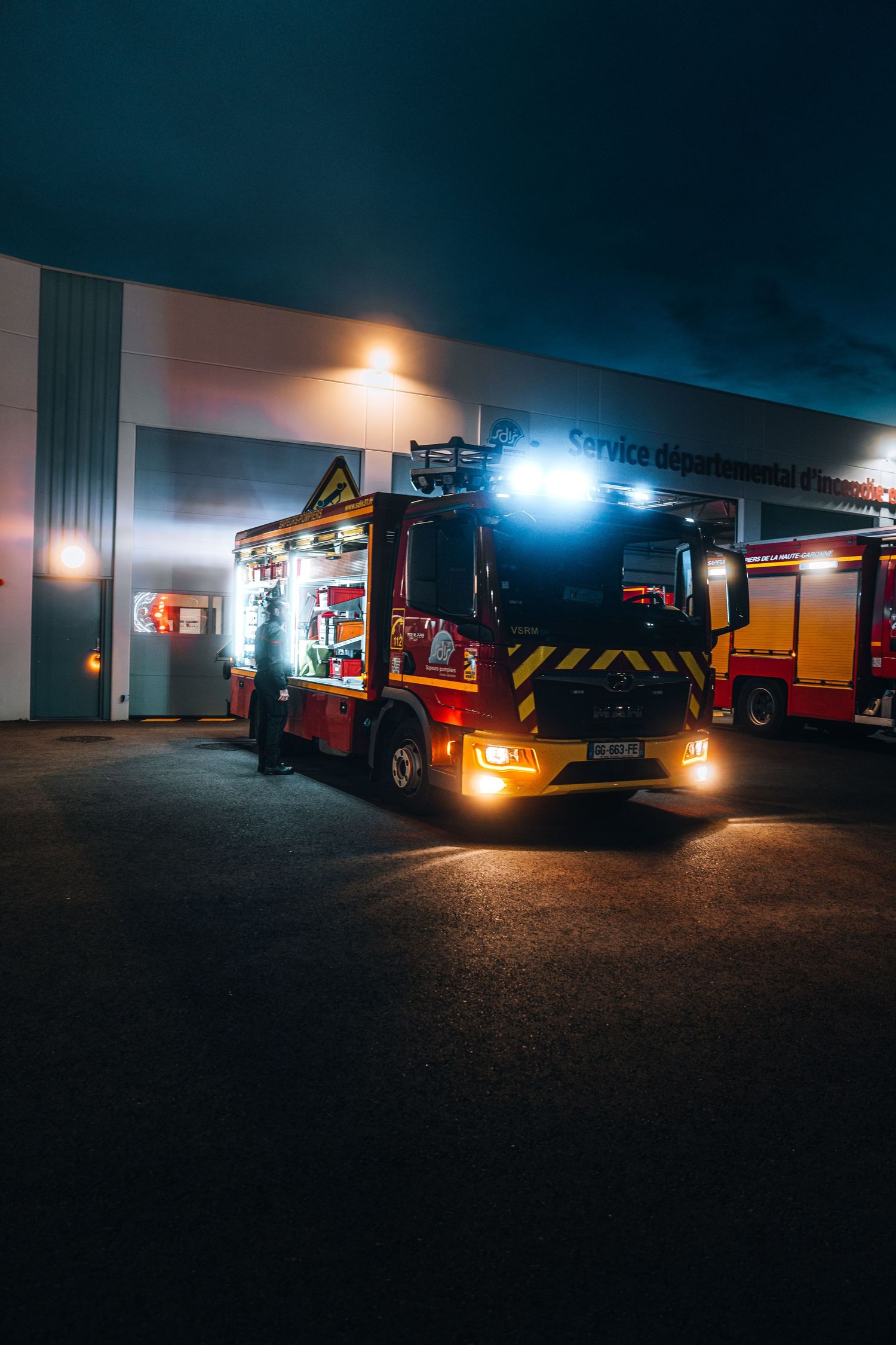 A red French fire truck with flashing lights parked outside a fire station at night.