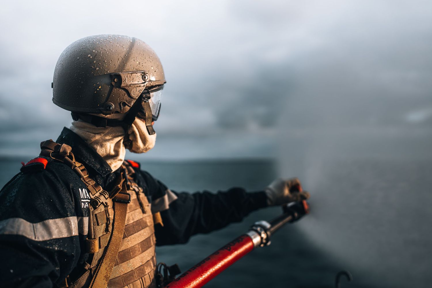 Navy soldier in tactical vest and helmet holds red equipment on a military ship at sea.