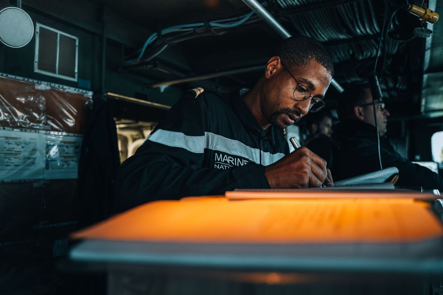 Marine nationale sailor writing notes under warm light inside a naval vessel during duty.