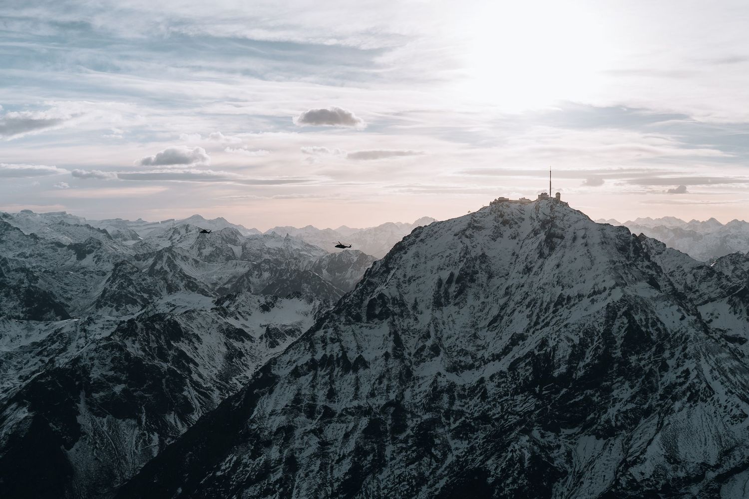 Panoramic aerial view of snow-covered Alpine mountains with a summit cross at dusk.