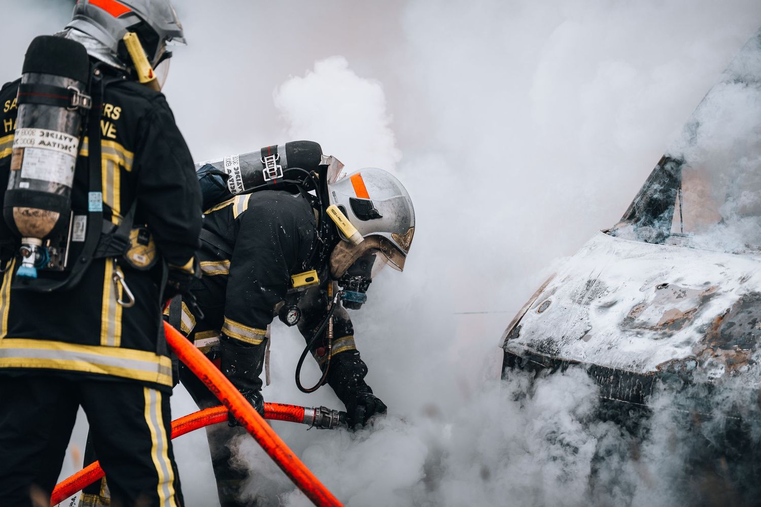 Firefighters in protective gear battle a smoking vehicle fire using hoses on a city street.