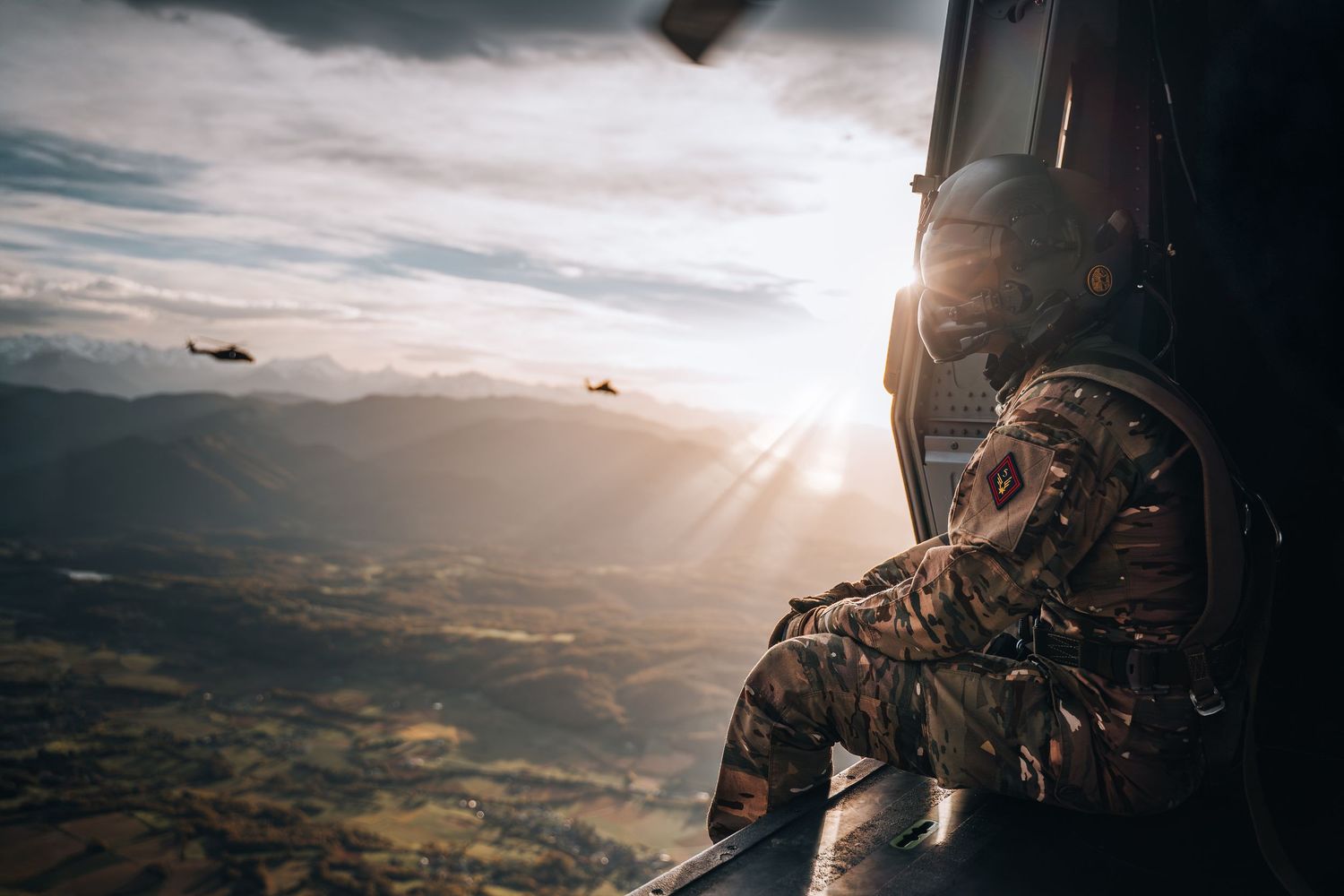 Military soldier in camouflage helmet observes aerial formation from open helicopter door over scenic mountains.