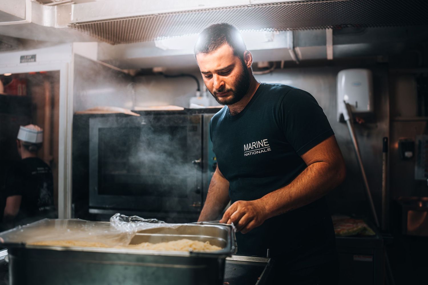 Chef in black uniform stirring large steaming pot in professional ship kitchen.