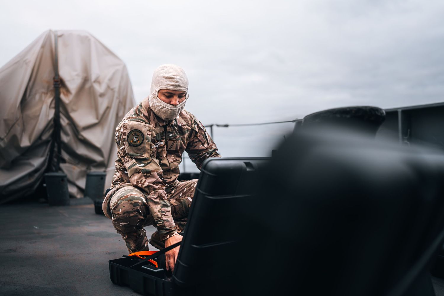 Camouflaged soldier crouches on naval vessel deck checking equipment in overcast conditions.