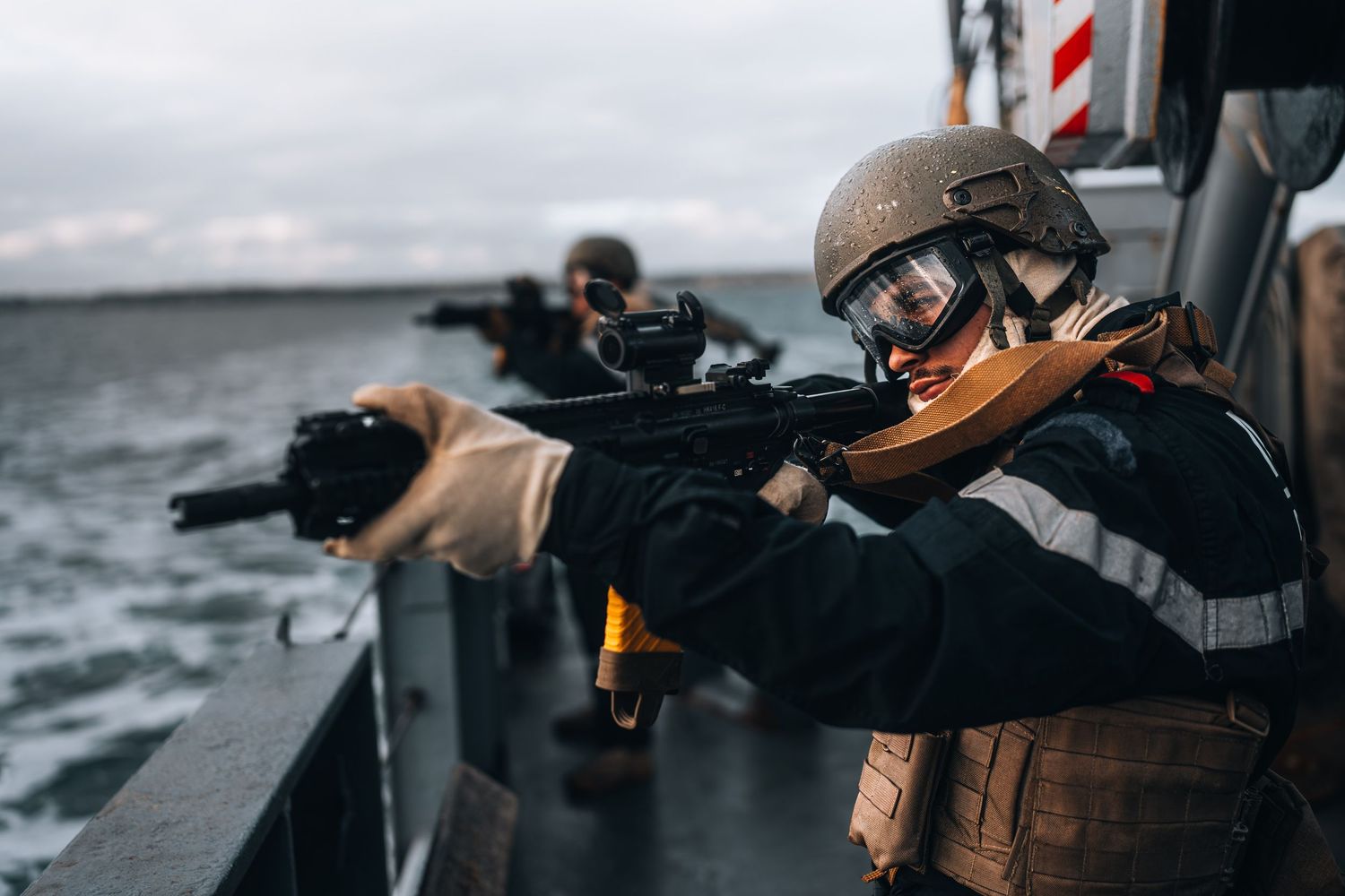 Two naval soldiers in tactical gear aim rifles from the deck of a military ship at sea.
