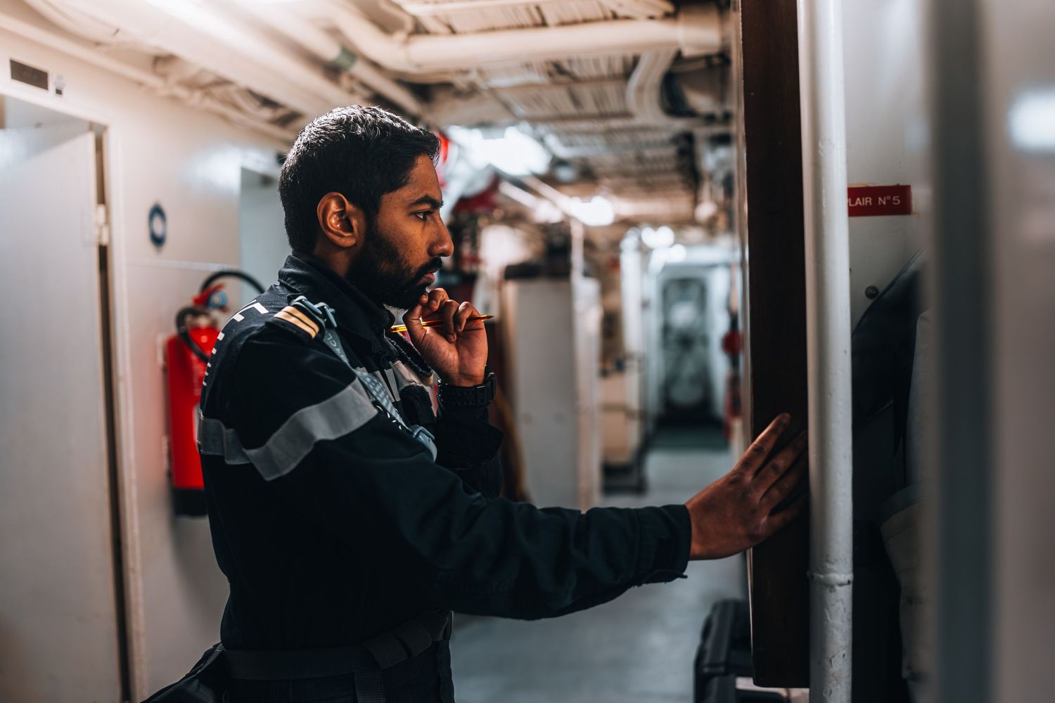 Military technician in uniform using radio while inspecting equipment panel in ship engine room.