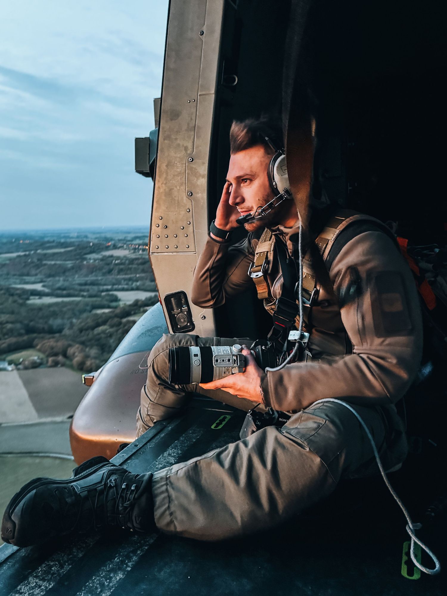 Aerial photographer in tactical gear sits in open helicopter door overlooking landscape.