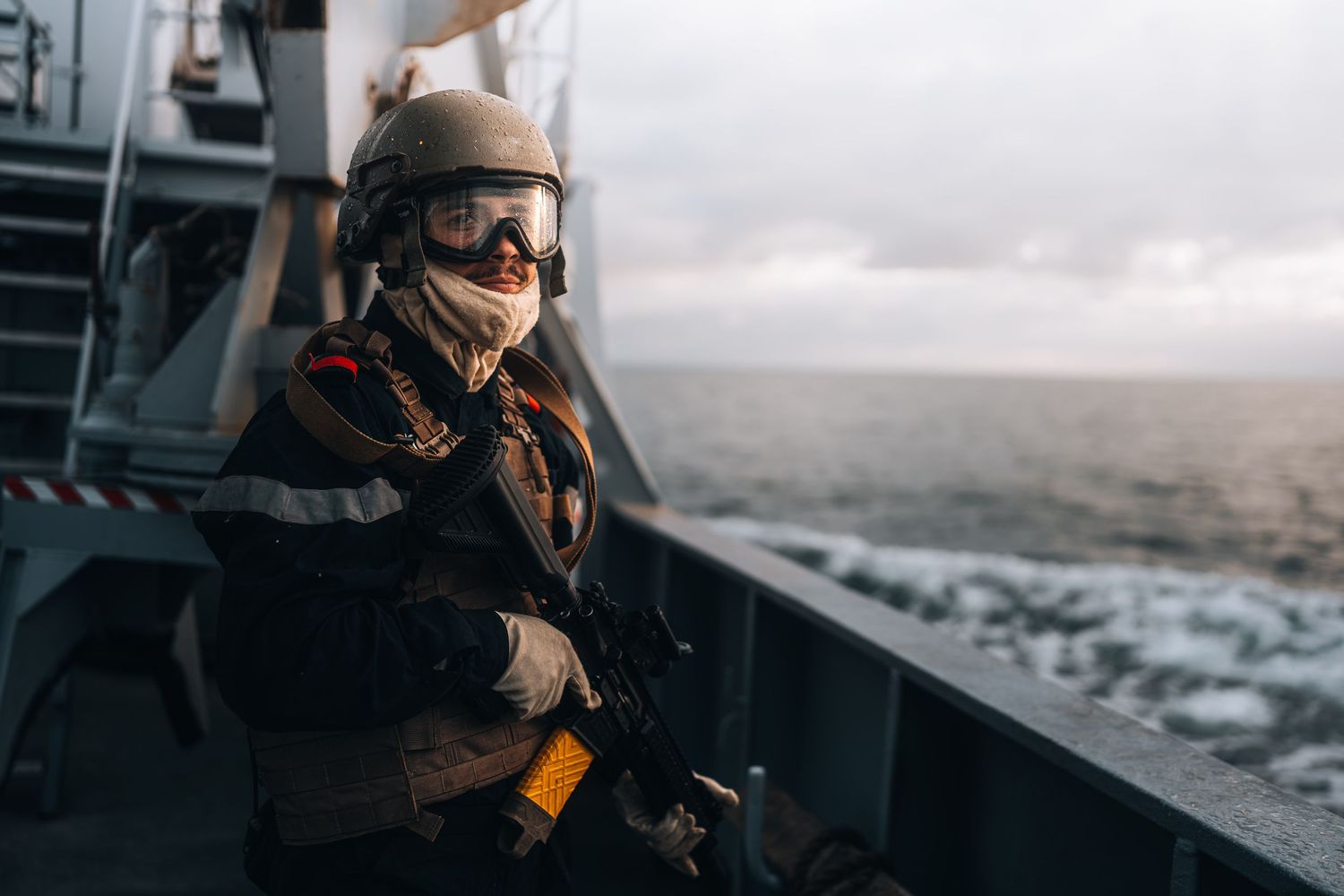 Armed soldier in tactical gear and helmet stands on a naval vessel deck overlooking the sea.