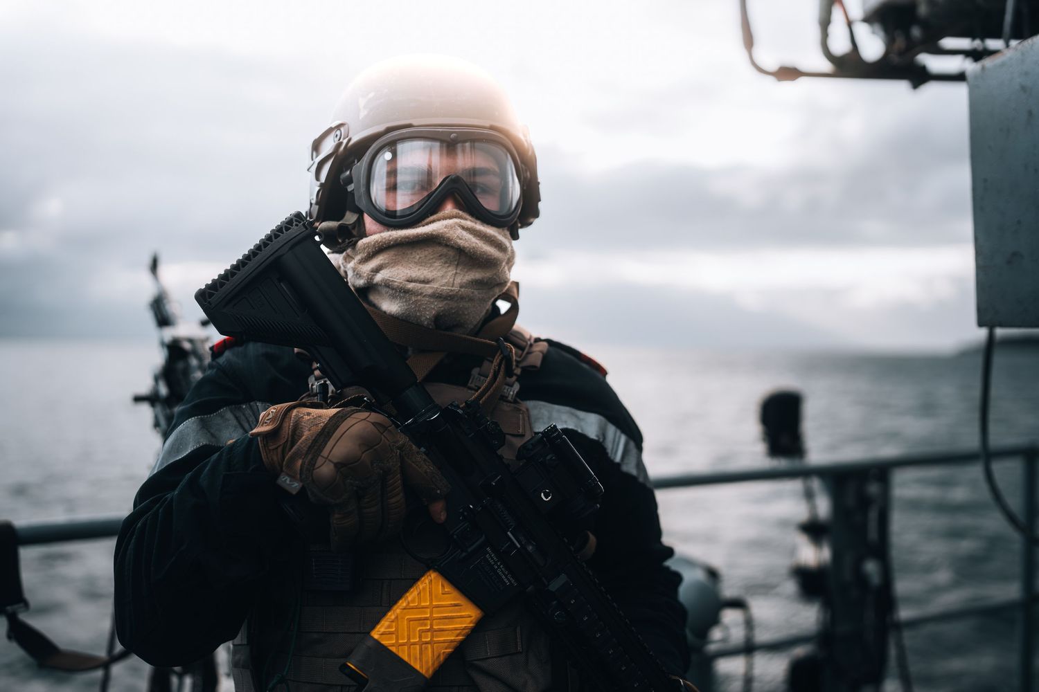 Armed navy soldier in tactical gear, helmet, and goggles stands on a military vessel at sea.