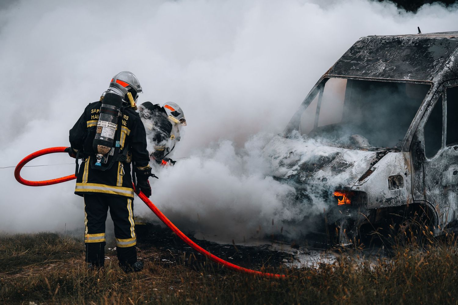 A firefighter in breathing apparatus uses a red hose to battle a heavily smoking vehicle fire outdoors.