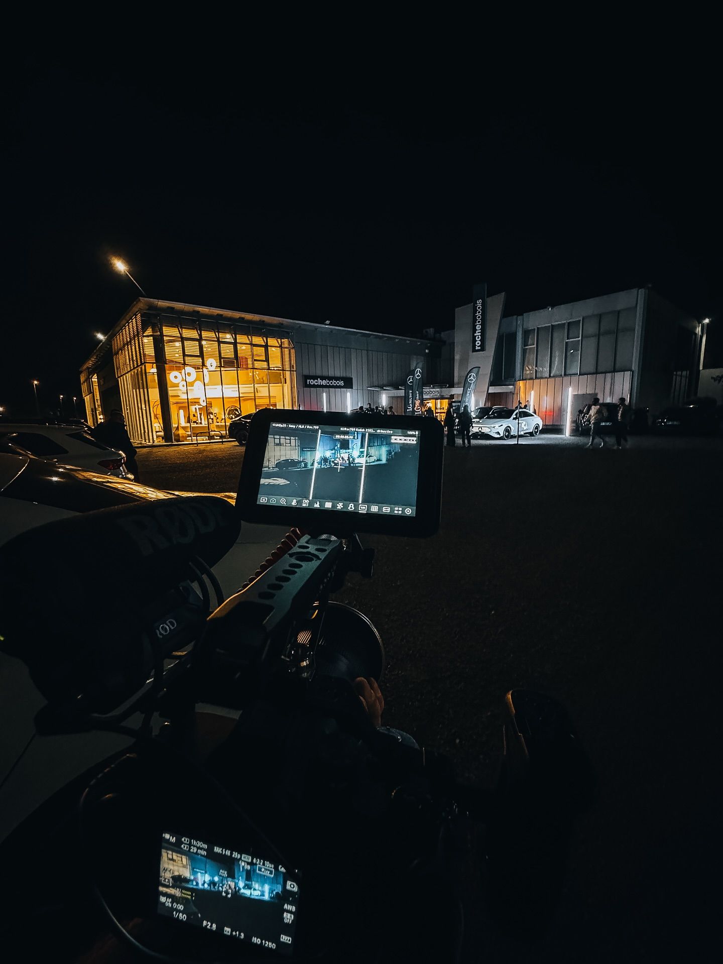 Nighttime behind-the-scenes camera setup filming an illuminated car dealership with luxury vehicles.