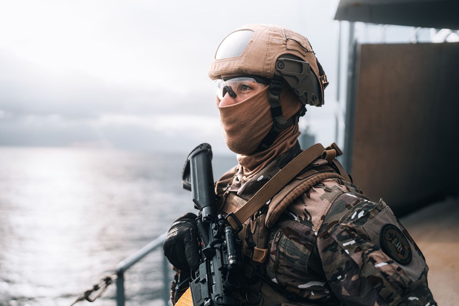Military soldier in camouflage gear and helmet holds rifle on naval vessel overlooking the sea.