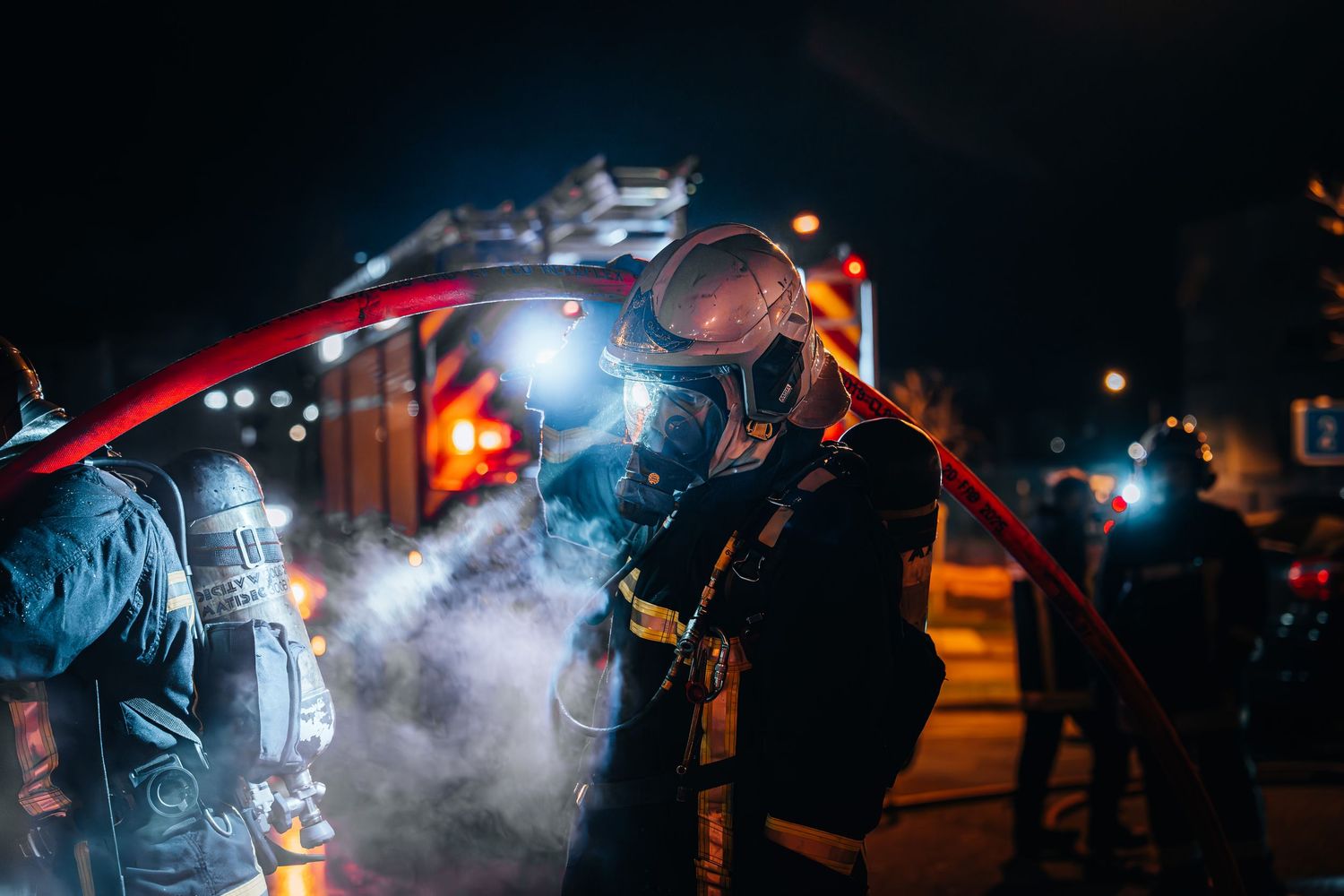 A firefighter in protective gear and breathing apparatus handles a red hose amid smoke at a nighttime emergency.