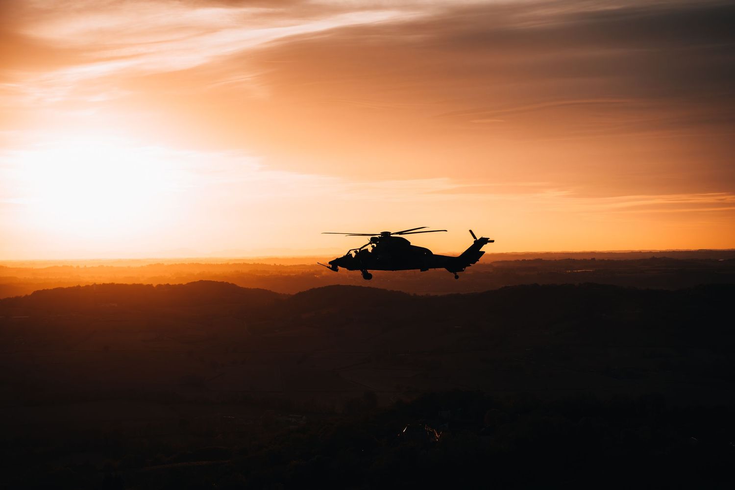 Military helicopter silhouetted against a dramatic orange sunset sky over rolling hills.