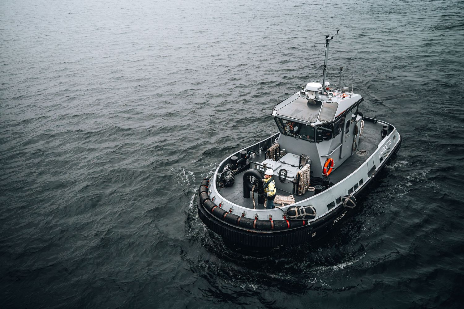 Military patrol boat with crew navigates calm gray harbor waters from aerial perspective.