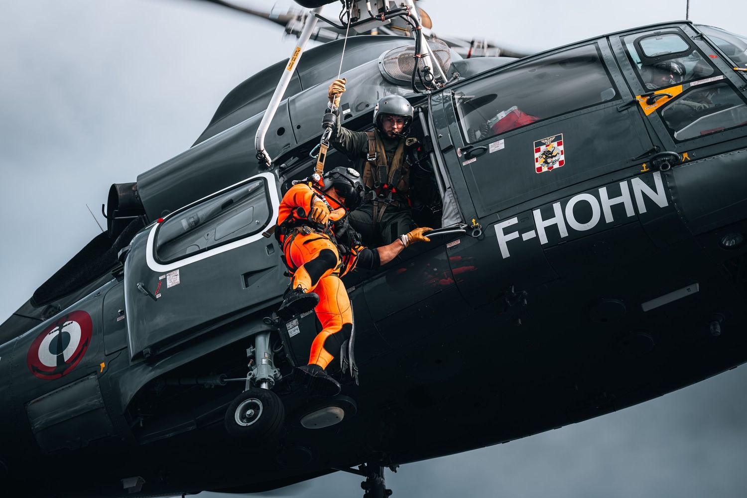 Rescue personnel in orange suit rappels from French military helicopter F-HOHN during operation.