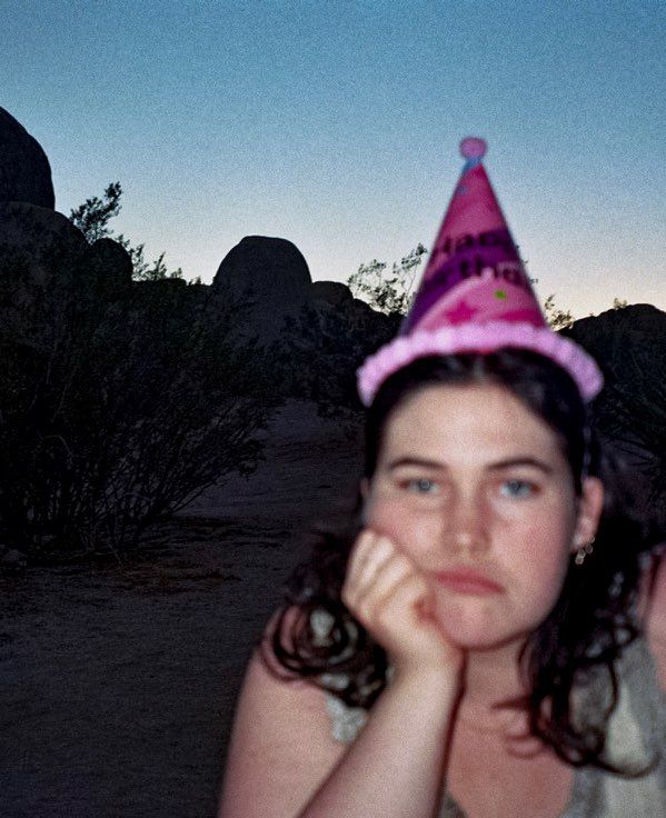 Young person wearing a pink birthday party hat poses outdoors at dusk with rocky landscape behind them.