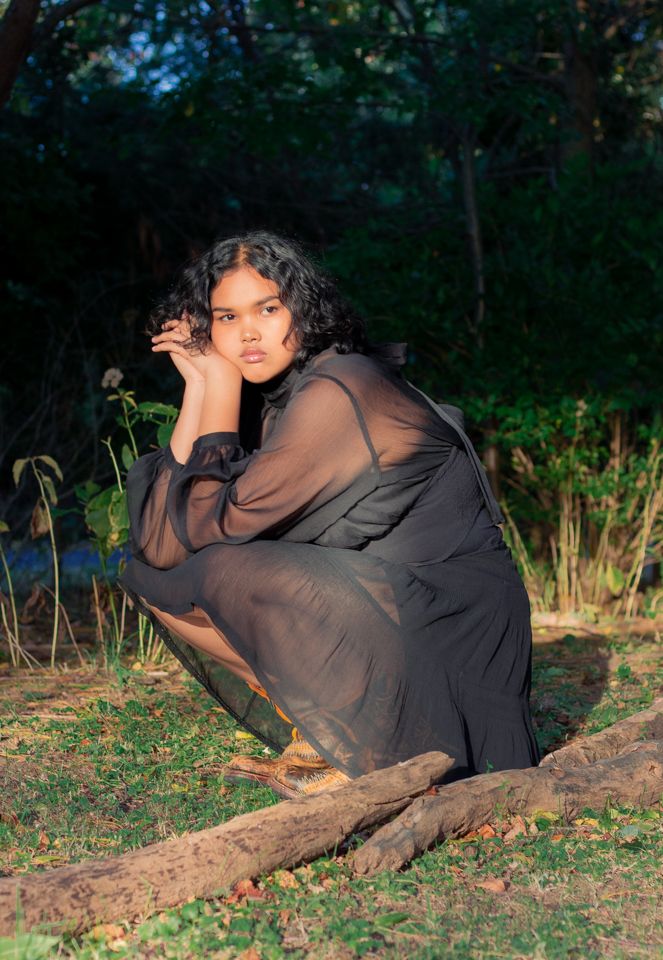 Woman in sheer black dress crouching near logs in a forest.