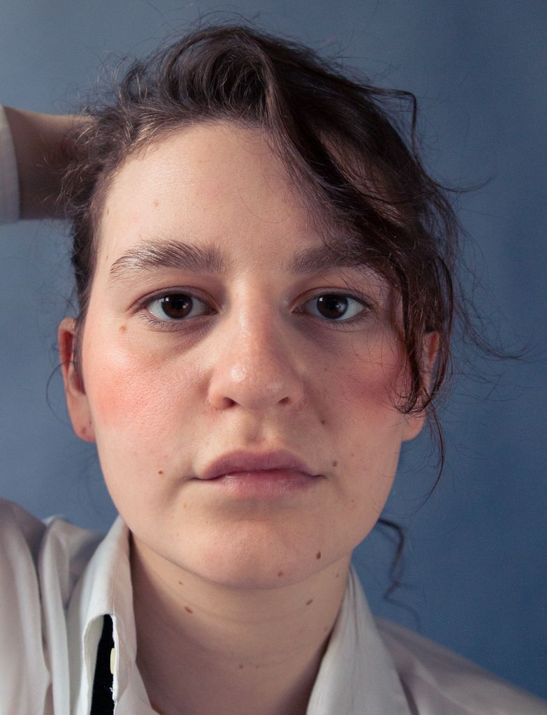 Young woman with rosy cheeks and dark hair held back by her hand in a white shirt.