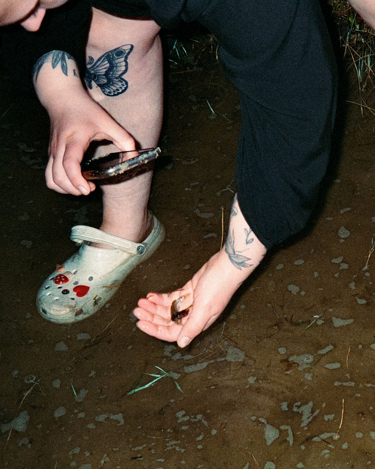 Close-up of tattooed legs and hands holding a small fish outdoors.