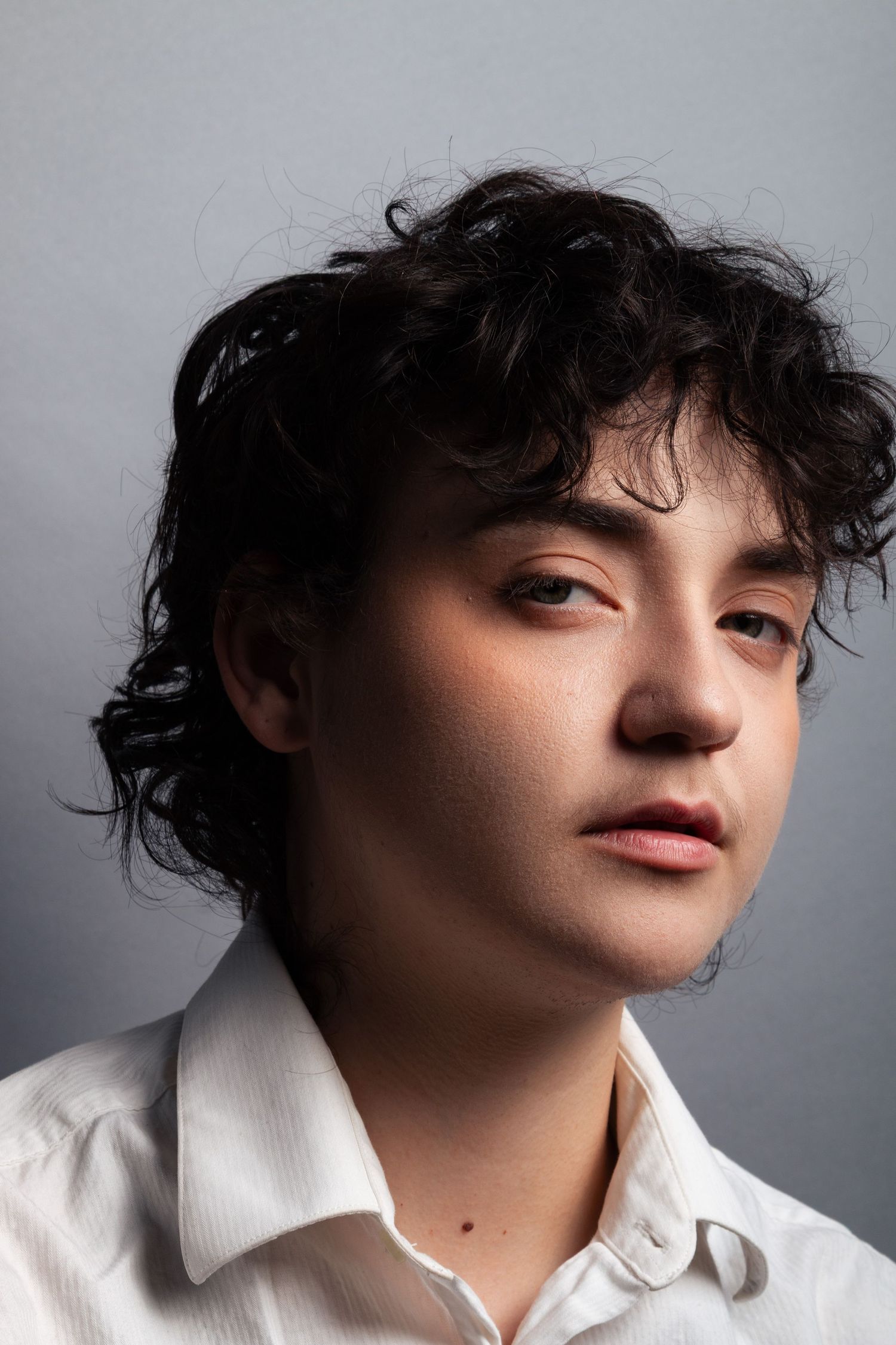 Young man with dark curly hair wearing white collared shirt, gazing upward against gray background.