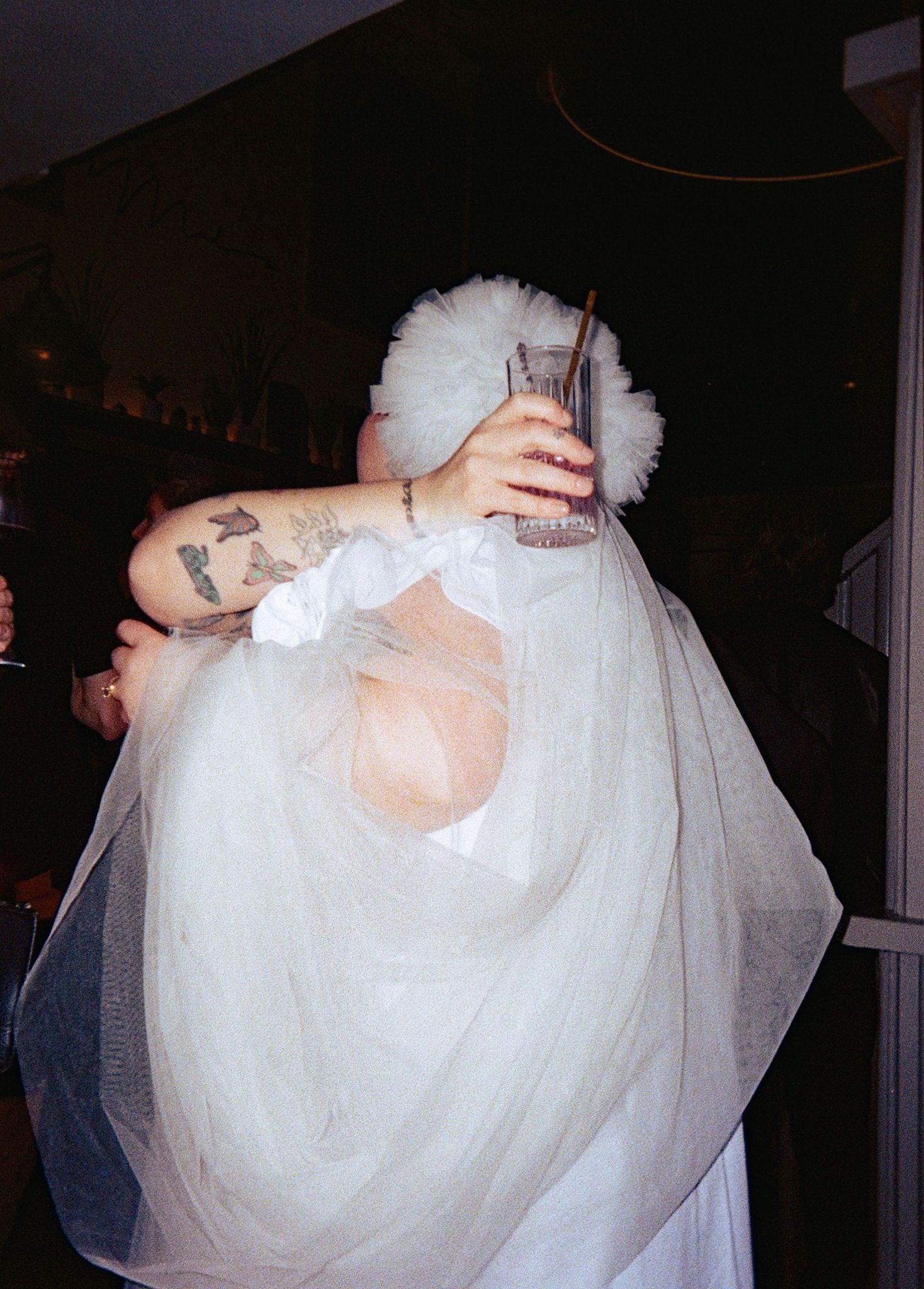 Person in white tulle dress and headpiece hugging tattooed person holding a drink at a wedding.