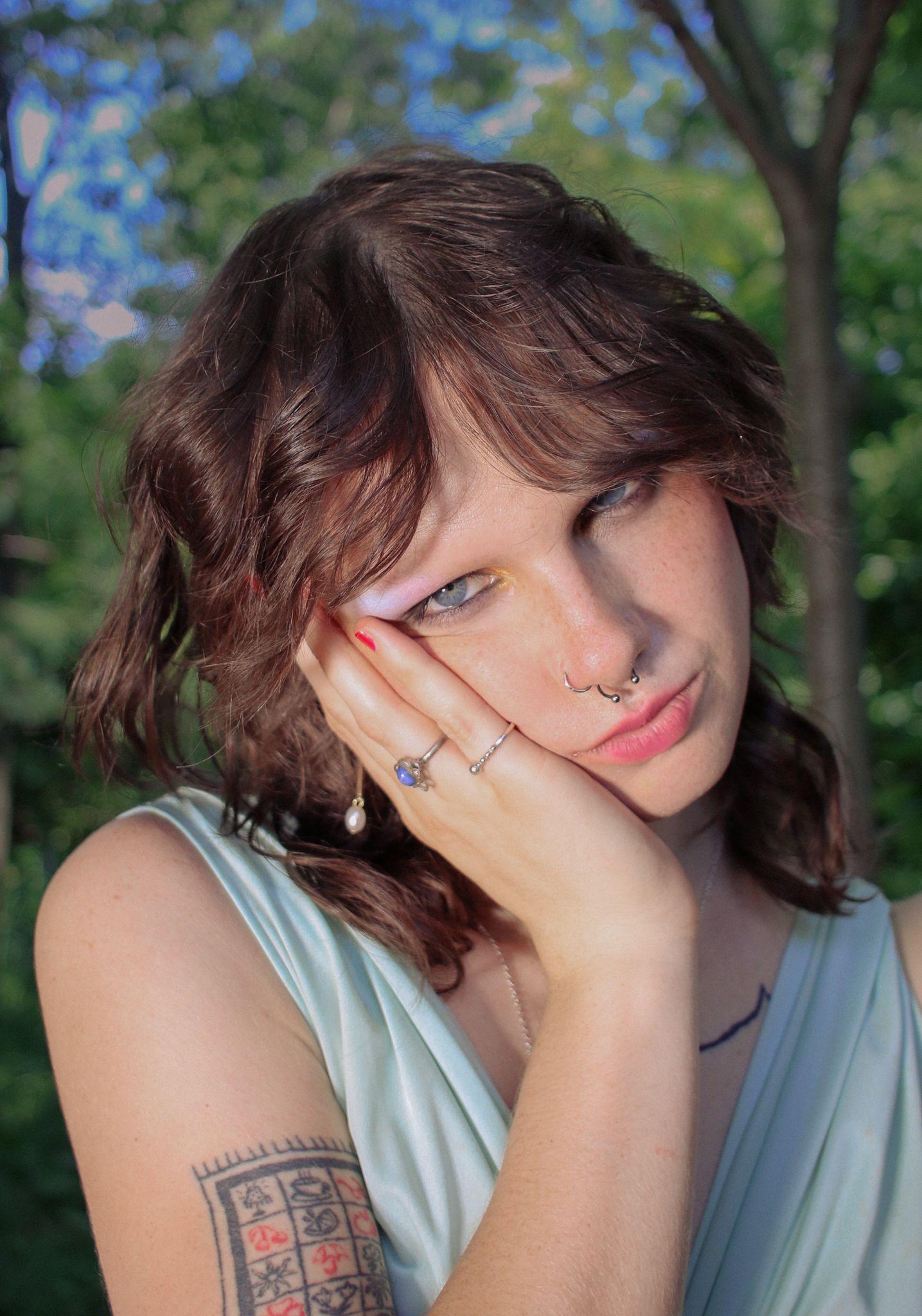 Young woman with wavy brown hair and nose ring resting cheek on hand outdoors among green trees.