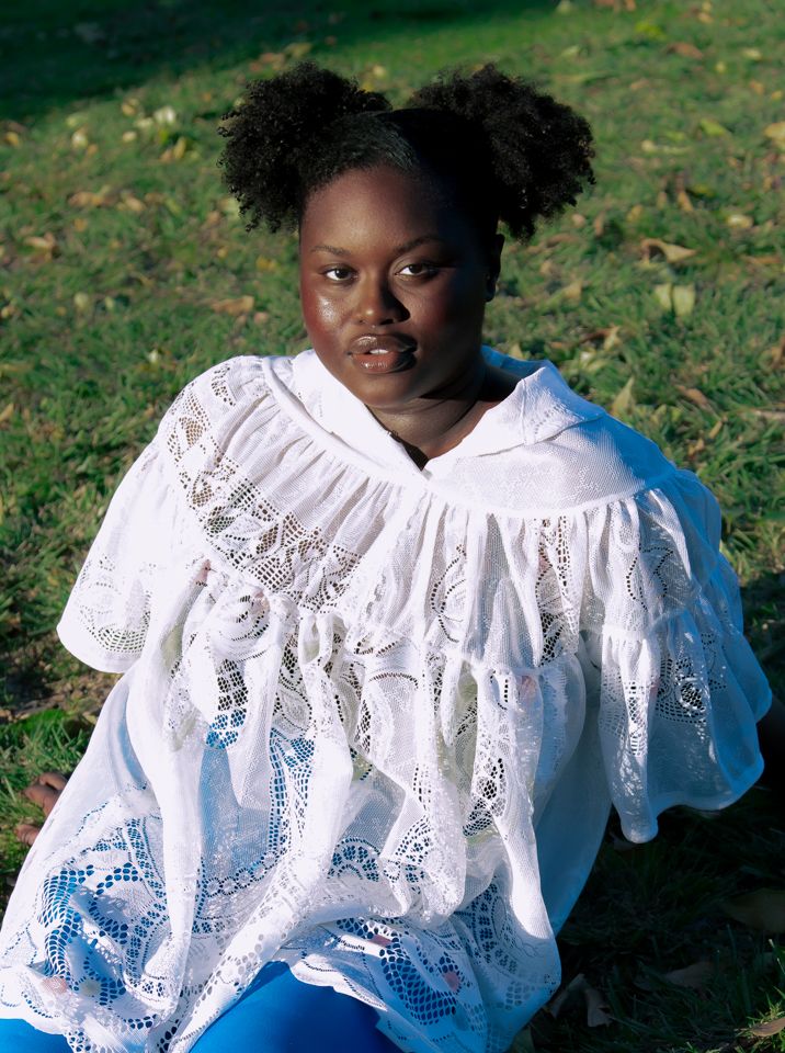Young woman with pigtails wearing white lace dress and blue tights sitting on green grass outdoors.