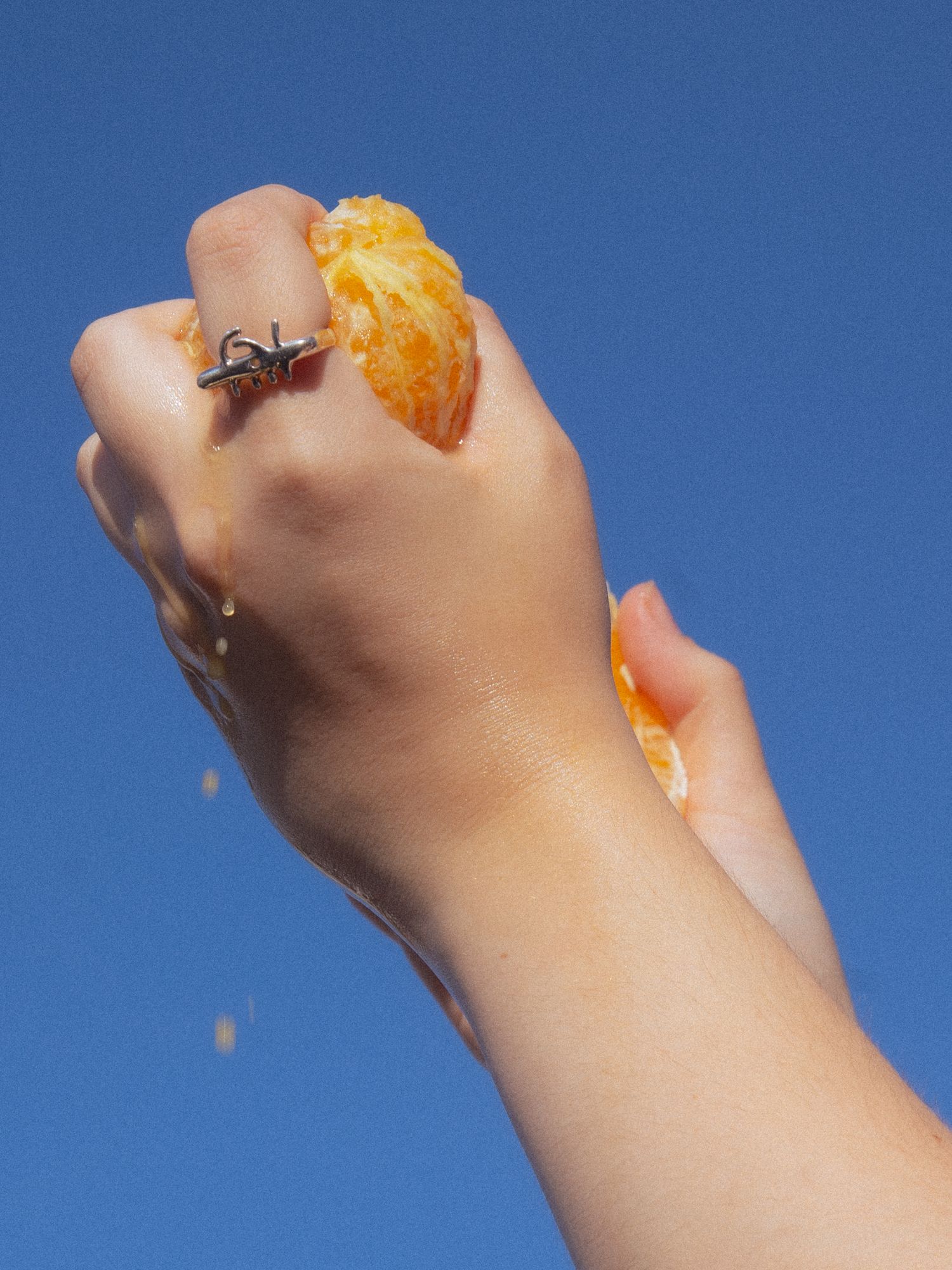 Hand with ring squeezing a juicy orange against a bright blue sky.
