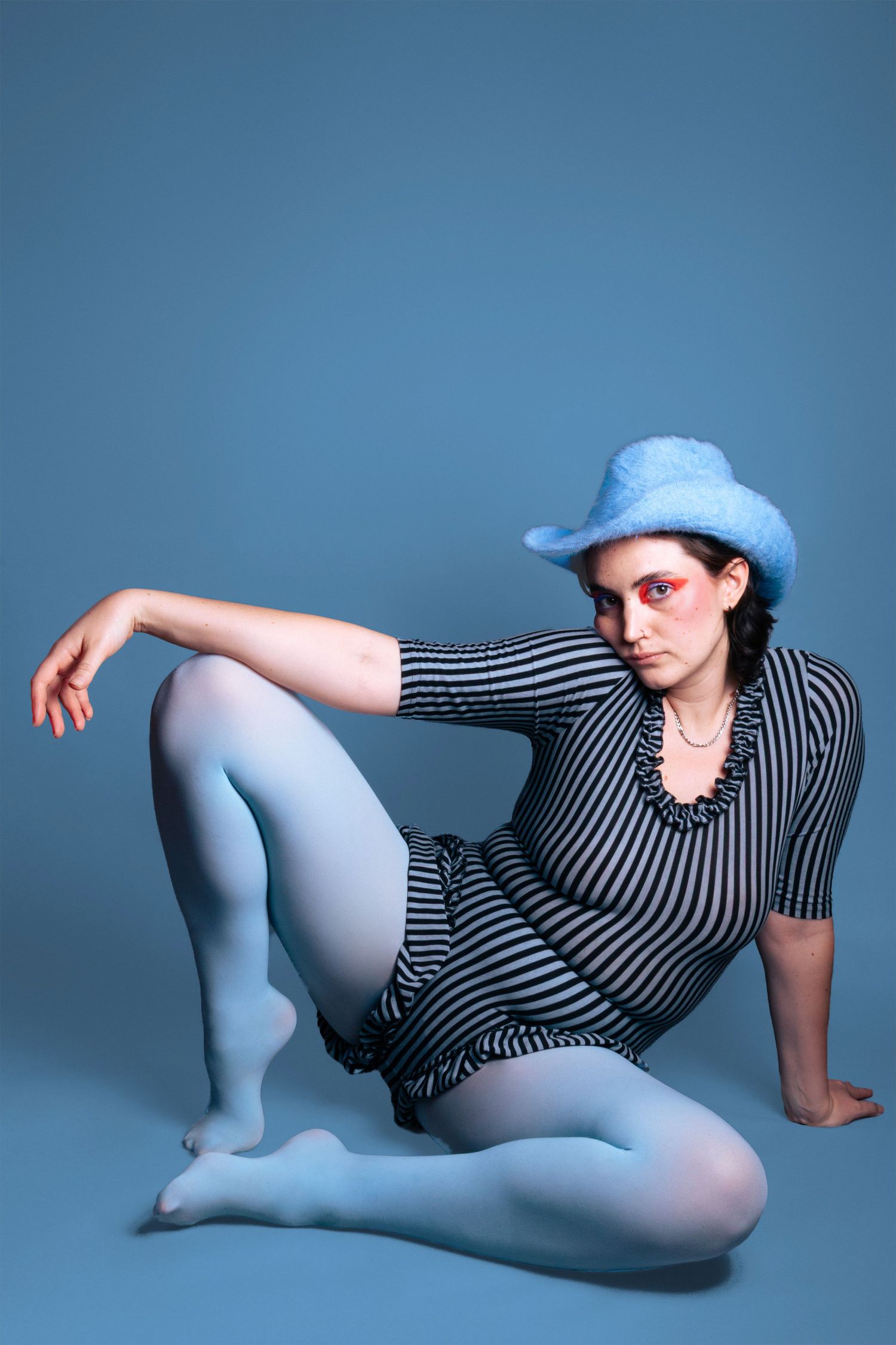 Woman in striped dress and blue fuzzy cowboy hat posing on blue backdrop.