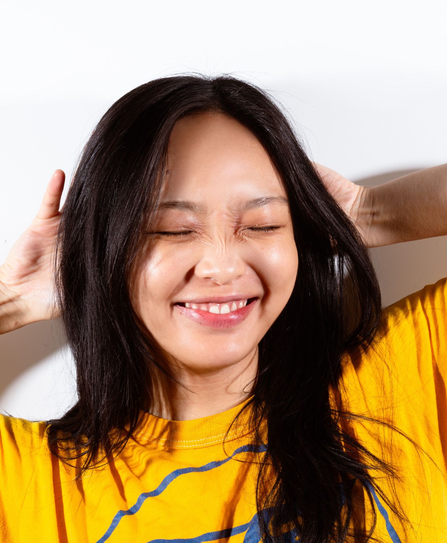 Happy woman in yellow shirt smiling with eyes closed, hands in long black hair, on white background.