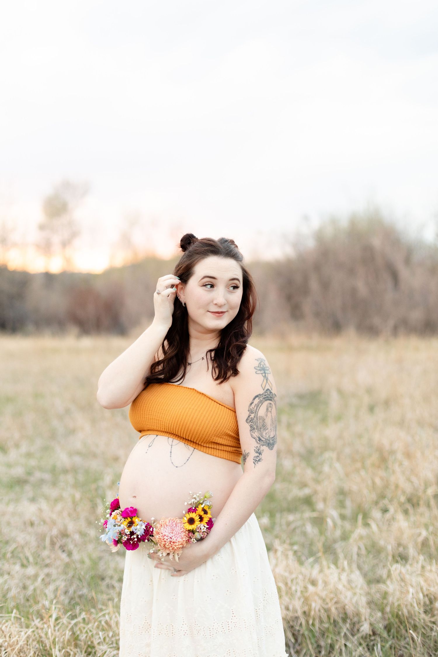 Pregnant woman in orange crop top and white skirt holds colorful flowers in a golden field at sunset.
